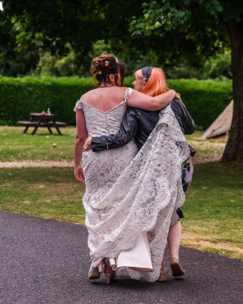 Jess is walking away from the camera. she has orange hair and a black headband and the bride has her arm around Jess' neck. The bride is wearing an off the shoulder lace dress with buttons all the way down the spine of the dress. The dress is ivory. They are in conversation as companions.