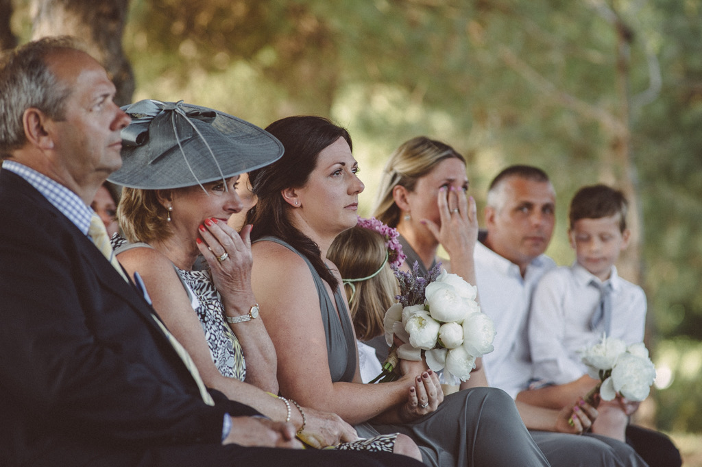 A Celebrant led wedding. Jess is the Celebrant. A row of wedding guests in a woodland. A man in a blazer to the fore. A woman in a grey hat in a saucer shape. A woman with dark brown long hair and a sleeveless bridesmaid dress in grey and a bunch of white peonies. next to her is a young girl facing away. Next to her is a woman with fair hair wiping her eyes and trying to stop herself Ching. and then there is a man with a boy on his lap looking to the camera and looking a bit lost. there is a lot of emotion