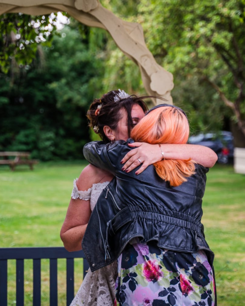 Jess is hugging a bride in a sleeveless lace dress. jess is wearing a floral dress in lilac and pink and has a leather jacket in black.