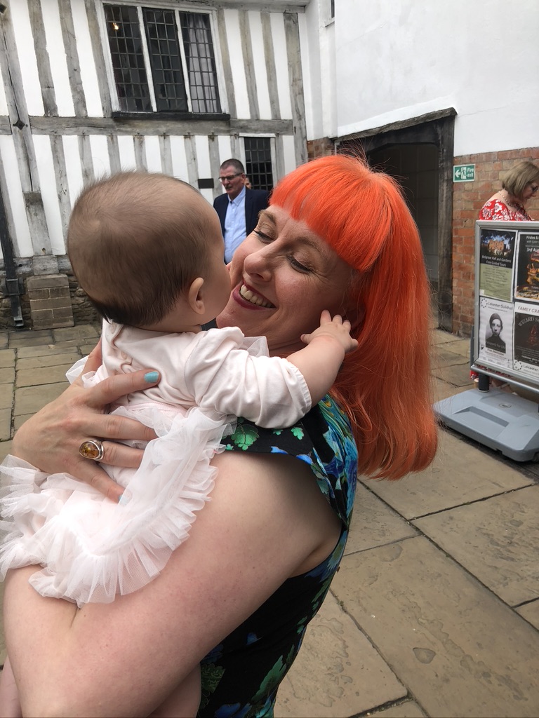 picture of Jess with bright orange hair, holding a baby. she has turquoise blue nails and an amber ring. the baby is holding her face and jess is smiling. there is a Tudor building in the background. it is white with grey wooden beams