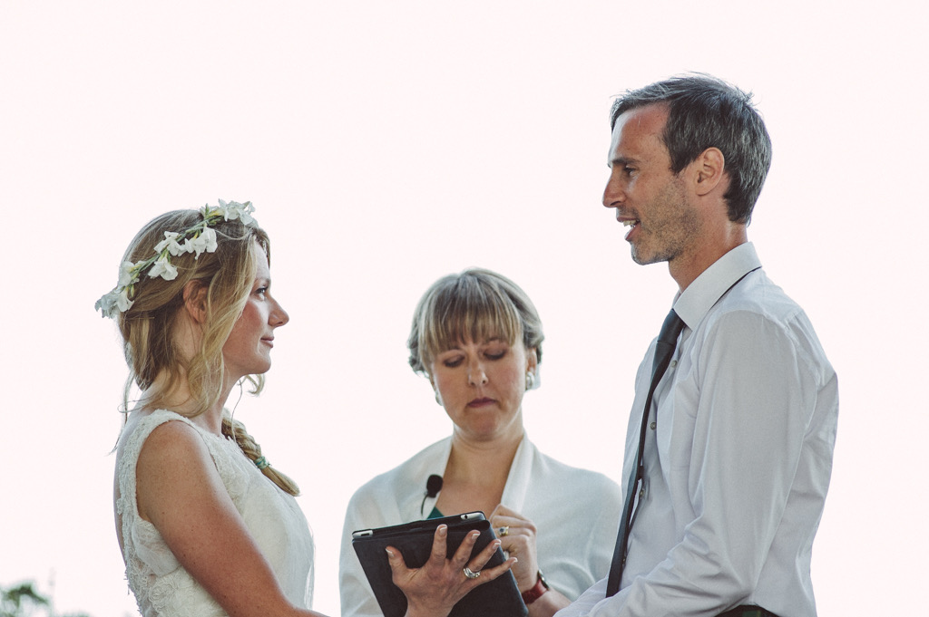 Jess is standing with an I pad. She is the Celebrant for a couple. a bride and a groom. The bride is on the left as you look at the photo and he is on the right. a female Celebrant is in between them. She has a fringe is holding a black I pad and has a lapel microphone. The bride is wearing an ivory sleeveless dress and has a coronet of flowers in her hair. the bride and groom are facing each other. They are saying their vows. they are outside