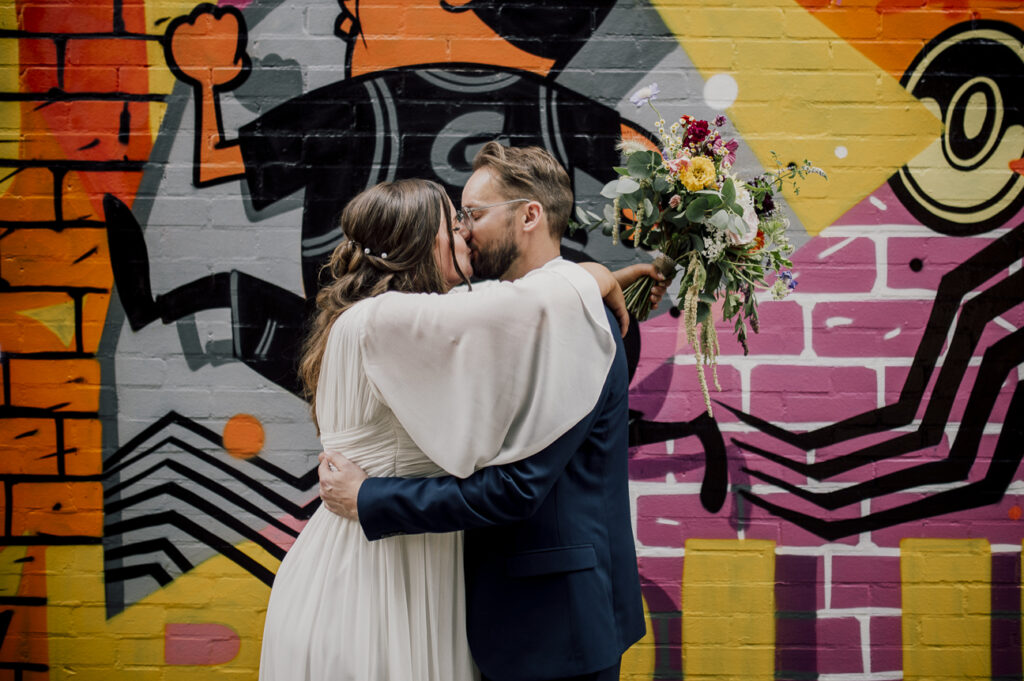pink and orange and yellow and black background with a bride and groom kissing . The bride has her arms around his neck. she has her hair braided loosely. her sleeves are floaty with white chiffon. She is on the left. She holds her wild flower bouquet behind his head in her left hand. the wall behind is graffiti art.