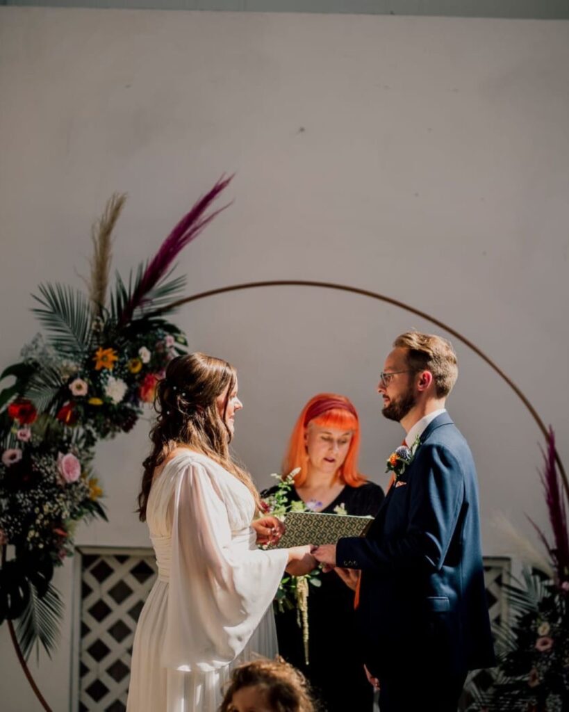 Jess stands in the centre of a circular backdrop with pampas grass. She is reading one of the 5 Great Wedding Readings. The light is magical and it falls on her bright orange hair. The couple are in the foreground. On the left is a bride with full floaty sleeves on a white dress to the right is the groom in a blue suit