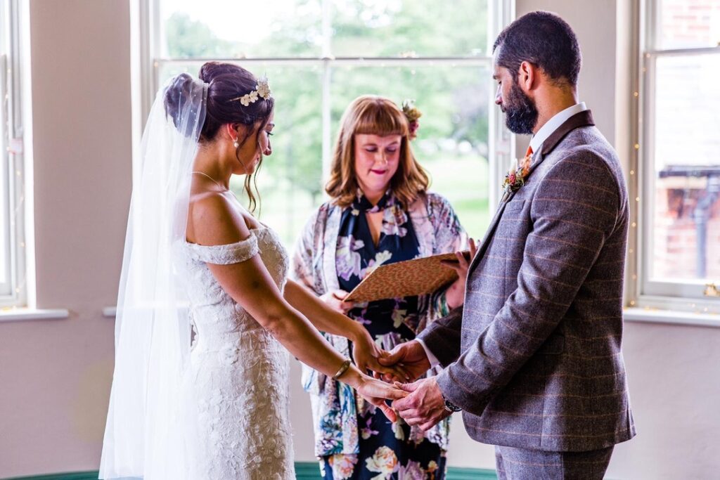 Brighton Wedding Celebrant Jess May is reading one of the 5 Great Wedding Readings jess stands in the middle. a tall man with a beard is on the right of the picture a bride is on the left. he is wearing a grey suit. she is wearing an off the shoulder white gown and a veil