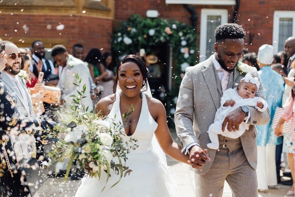 a Black couple. the Groom is on the right carrying a small baby. She has a bow in her hair. He has short hair. She is on the left and has a chic white dress with a halter neck and a large white bouquet. There is a confetti shot.