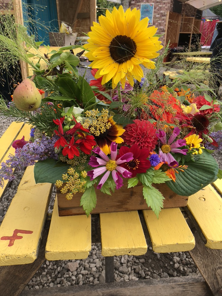 a yellow picnic table. with a floral arrangement with wild flowers from a British garden including red dahlia, pink Buddleia and red flowers. This illustrates an example of a funeral and how colourful they can be