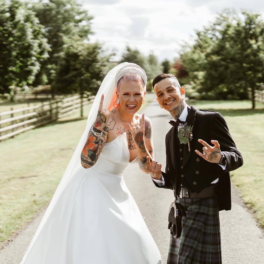 Tattooed couple. on the left is a woman with pink hair and a coronet like Queen Elizabeth the 1st. Both are fair skinned. she has pink eyebrows. she is sticking out her tongue in a rock and roll pose. she is covered in tattoos. they are orange and black. She is in a wedding dress and has a long veil . both are white. The groom is smaller than her, in a black tuxedo and a blue and green kilt with a sporran.
