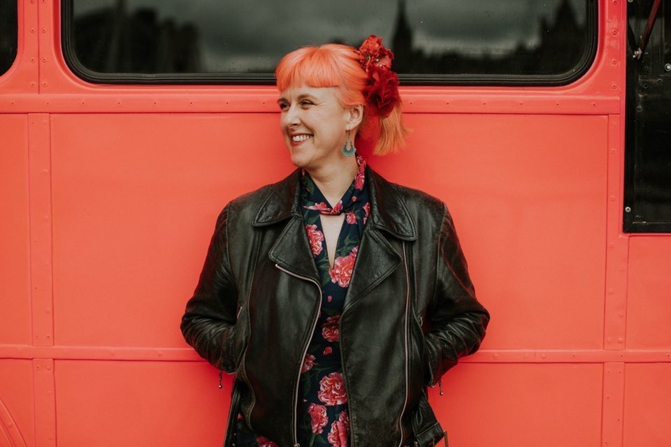 Jess is on an orange/pink background with her back to the pink/orange bus. she is wearing a leather jacket in black. she has red flowers in her hair. her hair matches the bus