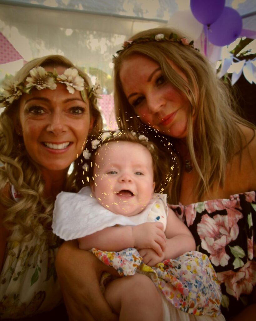 Baby Evelyn is being held between two women in their 30s. The women are Caucasian. They are smiling. on the left the woman has a white crown of flowers. The baby is smiling and also is wearing a crown of flowers. to her left as we look at the photo her Mum is also wearing a circle of flowers. they are tanned. They are in a marquee. it is summer. they both have long hair