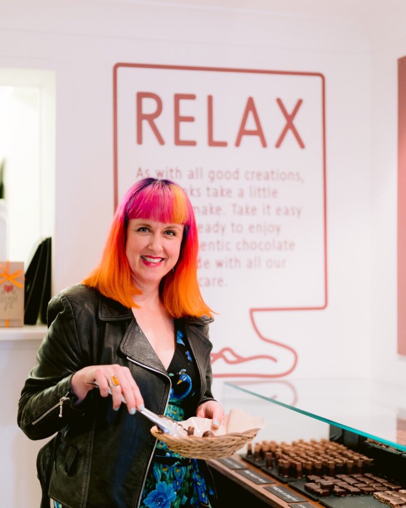 Jess is in a Chocolate shop choosing chocolates and picking them out with tongs. Behind her is a RELAX sign on a white wall. she is wearing a black leather jacket and has orange, pink and blue rainbow hair
