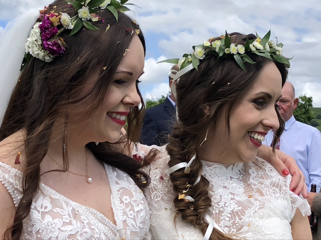 2 brides in ivory Boho dresses with applique lace. They have similar faces, with brown hair tied in a rustic informal style into curled braids. They are smiling and looking to their right. They look really happy