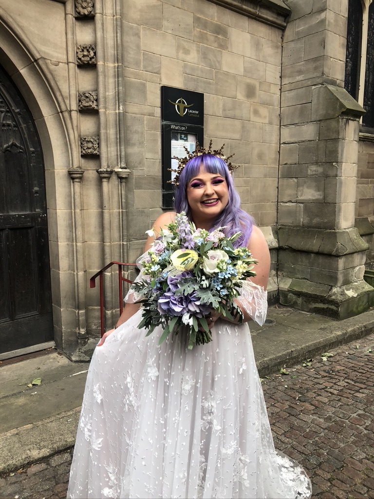 bride stood in front of an ancient Church. She has lilac hair. warm toned skin, a gold coronet and a bunch of lilac and blue flowers in front. her eye shadow is bright pink. her dress is floaty with lace and off white. it is 'off the shoulder'. She is smiling sweetly and looks really happy
