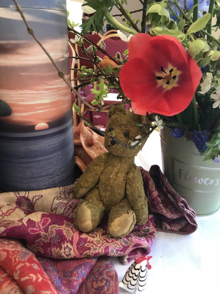A funeral image. A small brown teddy bear is sitting on a scarf. there is an urn with ashes. the urn has a sunset on it. There is a red poppy.The image is a photo of a funeral display