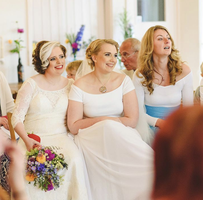 2 brides smiling in white dresses. one with a colourful bouquet. Out of the picture Jess May Brighton Wedding Celebrant is reading out one of the 5 Great Wedding Readings
