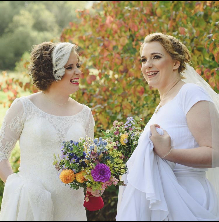 two brides in white wedding gowns, with an Autumnal background. one bride is holding her train and one is carrying a bouquet of wild flowers with pink and yellow