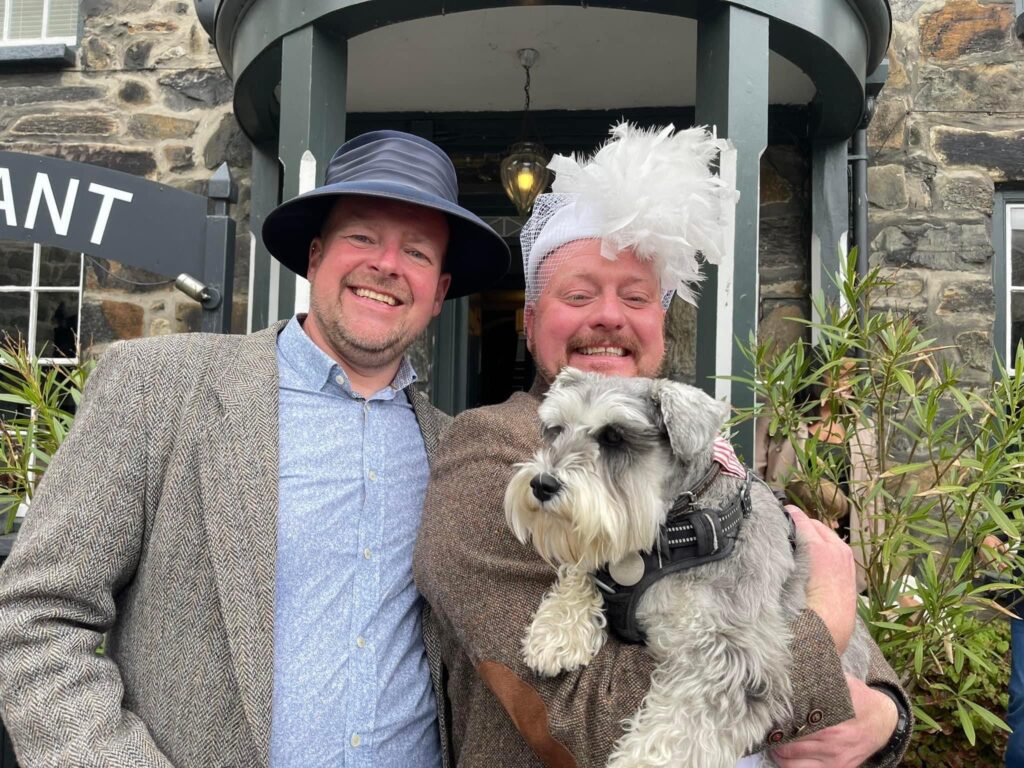 two men in ladies hats. They both have beards and sun tans making them look pink from the sun. They are in their 50s. the man on the right has a grey Schnauzer dog in his arms and a white feathered hat for a wedding. The man on the left has a blue shirt and a grey jacket and a blue straw hat