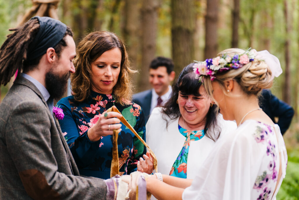 4 people doing a handfasting with ribbons. A woodland ceremony, spiritual but not religious. The groom is on the left and has dreadlocks and a bandana which is blue. then there is a woman wearing blue dress with pink flowers and brown wavy hair, then a woman in a white jacket and blue dress with black hair with a fringe and on the right is the bride in a white dress with a lilac coronet and a bun. she has lilac embroidery on her dress
