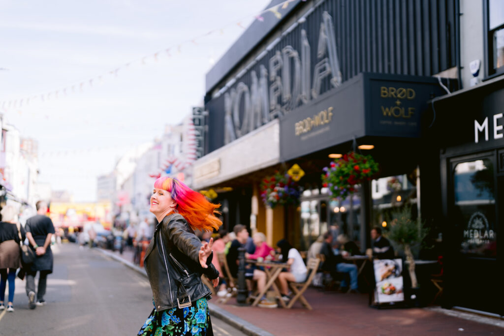 Jess May, Brighton-based Celebrant is whirling around. She is wearing a black jacket and a blue and black dress. She is in front of Komedia in Brighton. The background is black. Her hair is multi-coloured, she is swirling around and facing to the left