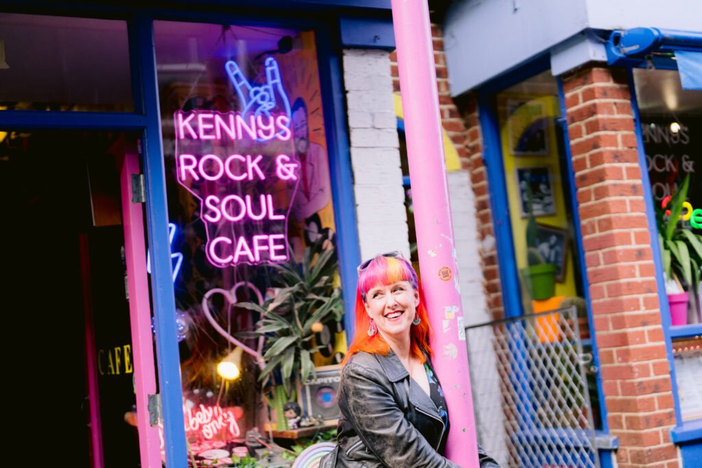 Jess May, Brighton-based Celebrant  is holding a purple column with both arms. She is standing in front of a pink sign for Kenny's Rock & Soul Cafe which is neon. the rest of the image is blue. She has sunglasses on top of her head. ear rings. a pink fringe with a yellow streak and orange hair. her jacket is black