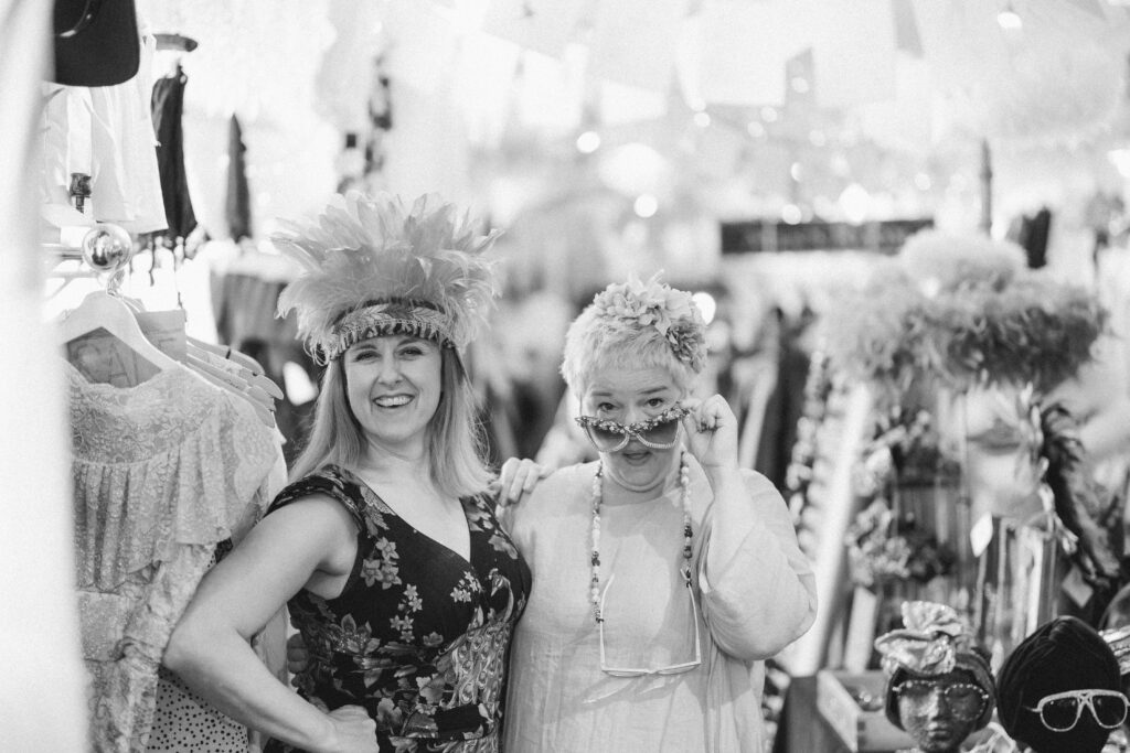 Black and white photo with a middle aged Caucasian woman. Jess is wearing a sleeveless dress and a big feather head dress. she has her arm around her companion who has short hair and is also caucasian. This woman is peeking over her glasses in a gesture of fun