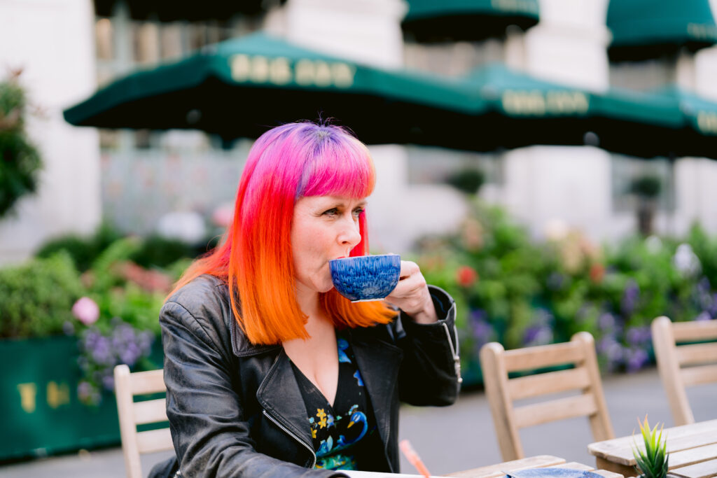 Jess May, Brighton-based Celebrant is drinking a cup of coffee in Brighton in front of The Ivy Hotel. She is drinking from a blue ceramic tea cup. The cup is large. She is wearing a black jacket made from something that looks like leather. She has rainbow coloured hair which is blue at the parting and then has a bright pink fringe and grading from pink to orange. her hair is shoulder length. She is sipping coffee. The Awning behind her is dark green with The Ivy written in bold Gold lettering