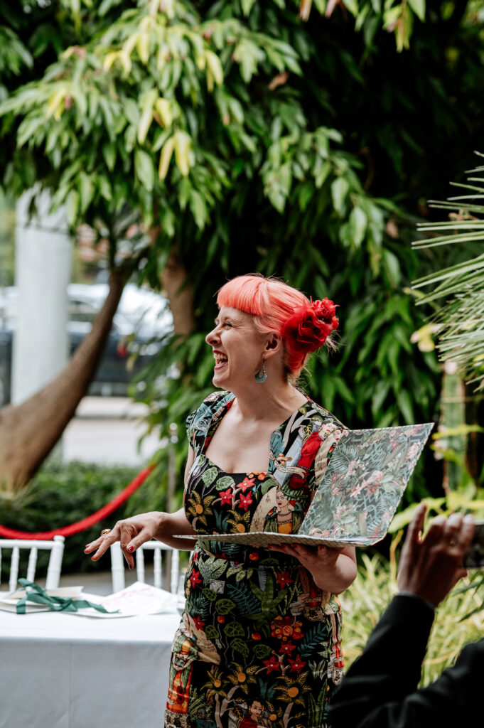 Jess laughing while holding a folder with a jungle pattern and wearing a Frida Khalo dress, she has orange hair and a red flower and is standing in a hot house. She is reading one of the 5 Great Wedding Readings.