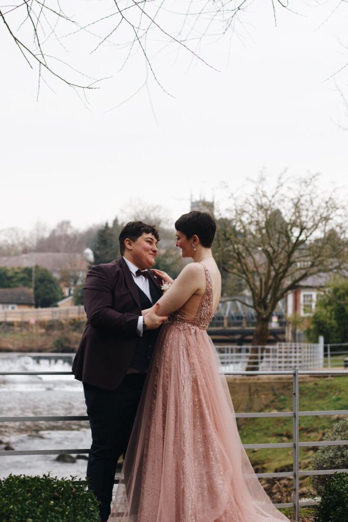 A lesbian couple stand in an embrace. they have a backdrop of a river, a tree and a church tower. One bride is wearing a burgundy suit with a bow tie and a white shirt, the other is wearing a backless elegant gown in blush pink with sequins. both have dark cropped hair