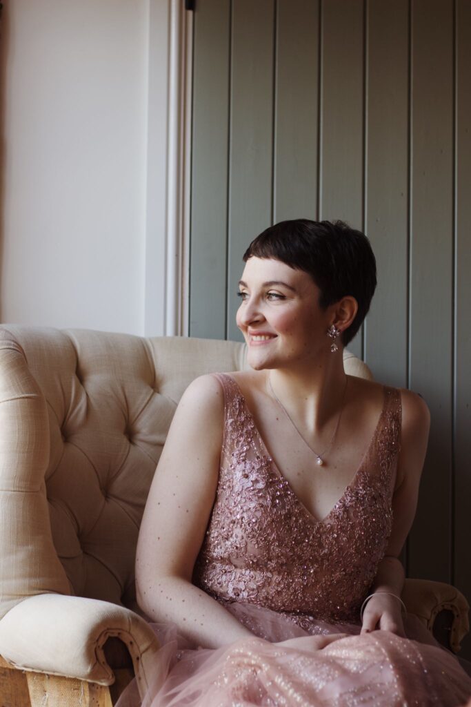A beautiful image of a bride with cropped dark hair looking like Audrey Hepburn.  She is in a blush pink sequinned dress. the dress is sleeveless. She is looking out of the window smiling whimsically. She has porcelain skin and looks calm. she does not have wedding day anxiety