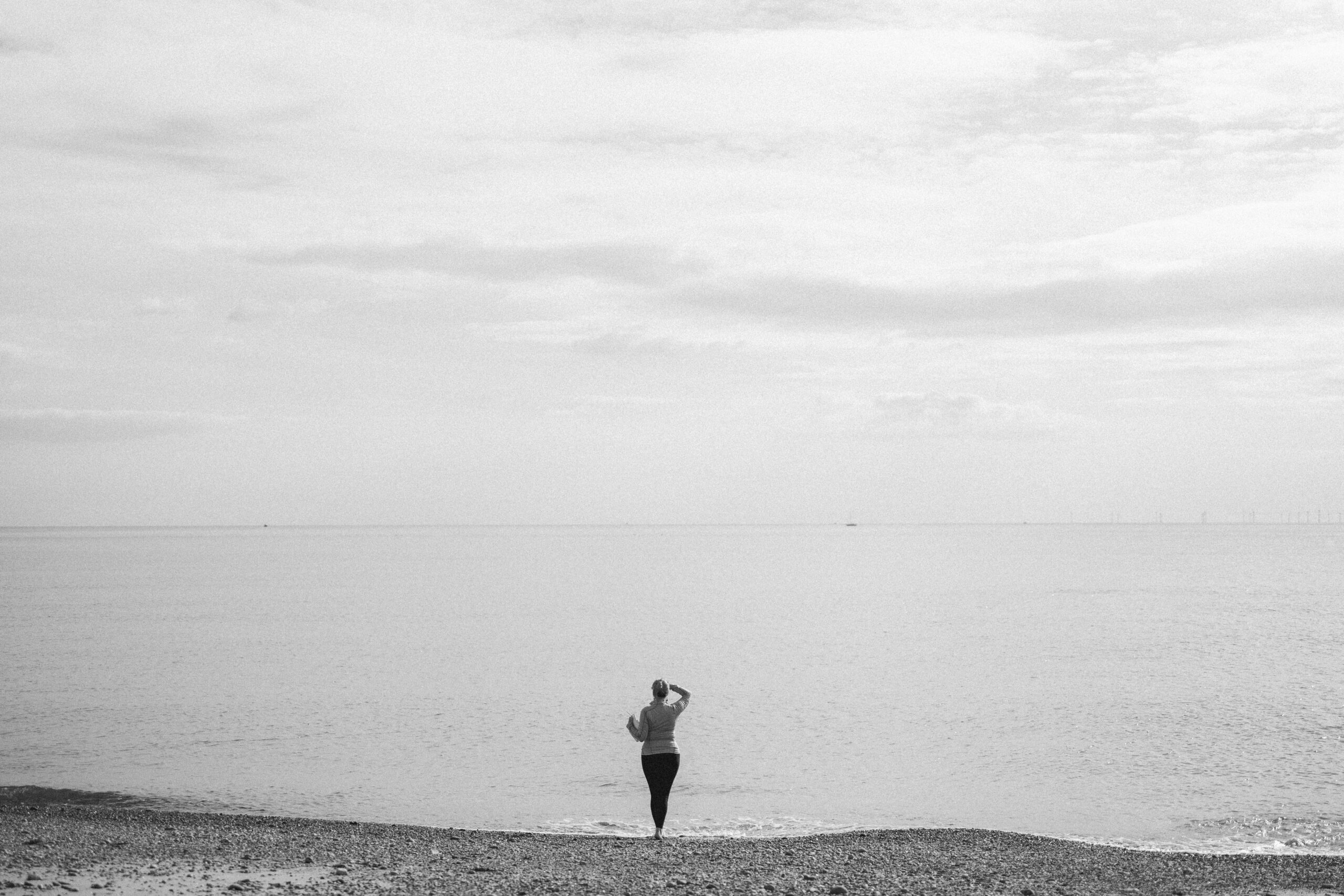 A grey scale, black and white photo of a beach. A woman is standing looking out to sea and has her hand up to shield her eyes. This picture captures the essence of how it feels to see new horizons. Jess May has just discovered her Vocation as a Celebrant