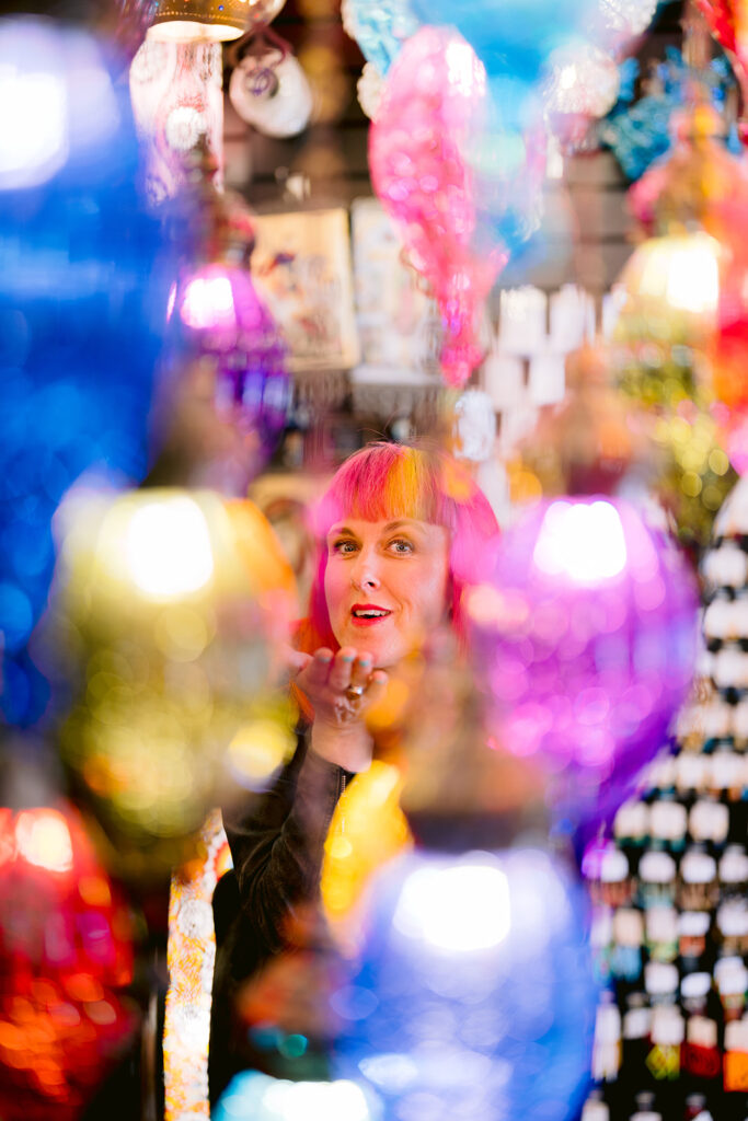 Jess is sitting behind many hanging coloured lanterns. She is blowing a kiss through them. it is a whimsical image