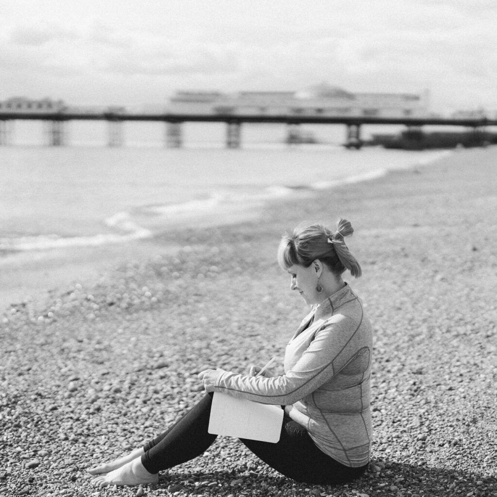 black and white photo. Jess is sitting on Brighton Beach. she has the pier behind her. She is writing a story other lap. it is a casual pose