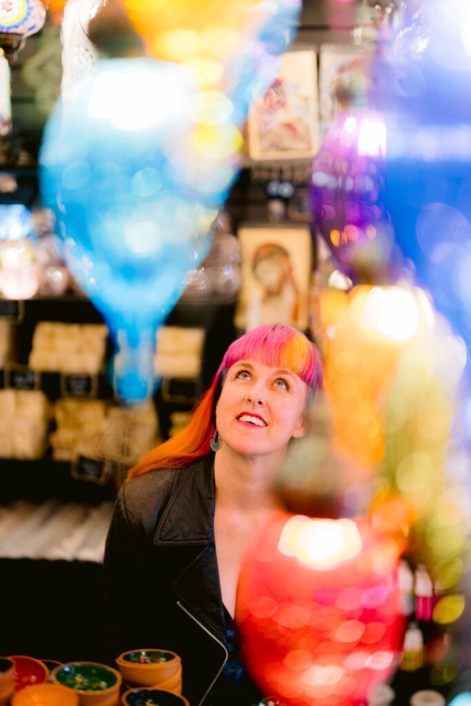 Jess May, Brighton-based Celebrant is sitting looking up surrounded by coloured glass hanging lanterns. The lanterns are turquoise, yellow and pink. She is smiling upwards. She is happy. She is wearing a black leather jacket and has a bright pink fringe with a yellow streak and the rest of her hair is bright orange.