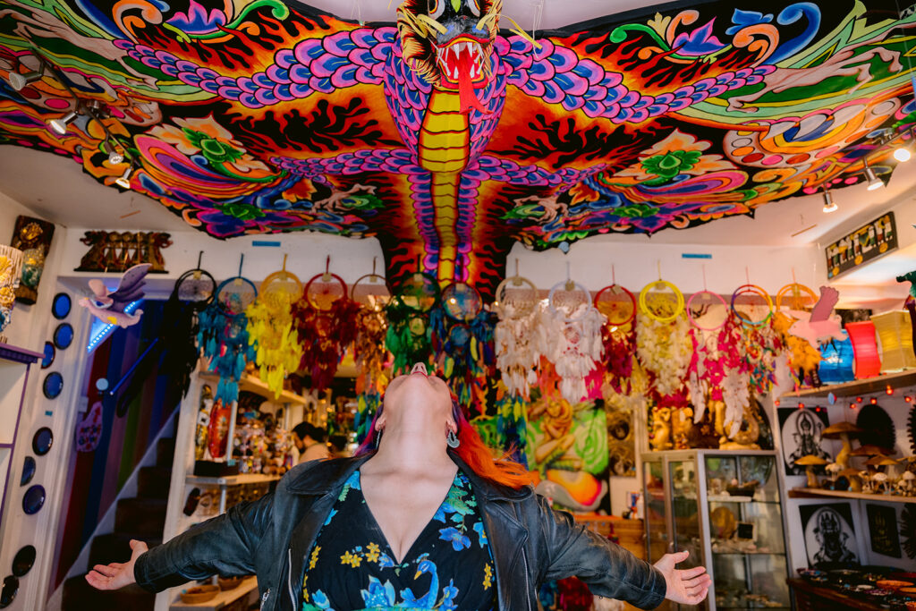 Jess May, Brighton-based Celebrant is standing with arms outstretched replicating the shape of a huge vibrantly coloured dragon shaped kite. There are rows of feather dream catchers behind her. she is posing in a gesture of liberation and freedom