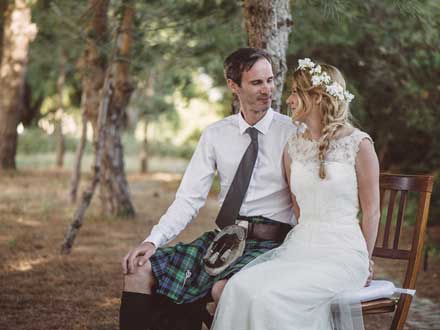 A bride and Groom sit next to each other. They are in love. She wears a white sleeveless floor length lace dress. he is in a kilt and tie with a white shirt. his kilts blue and green. They are gazing into each other's eyes. She has a coronet of white flowers and a plait. She is to his right as we look at the photograph. they are in a woodland in Summer